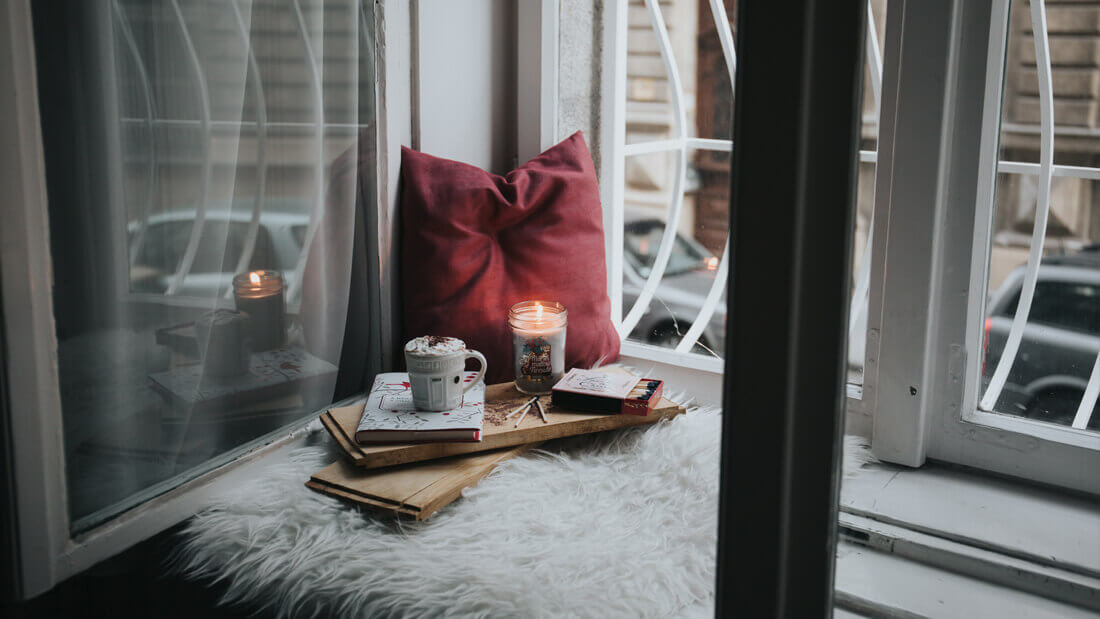 Hot chocolate and candles on a tray table and a red pillow placed near a window.
