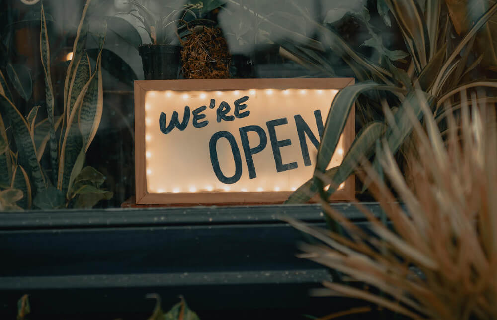 We're open sign against a window and indoor plants.